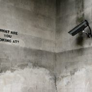 Caméra de surveillance observant un message écrit sur un mur, et qui dit : "What are you looking at?"