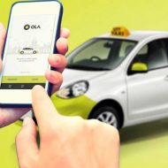 Ola : l'application VTC lancée sur un smartphone devant une voiture de la firme.