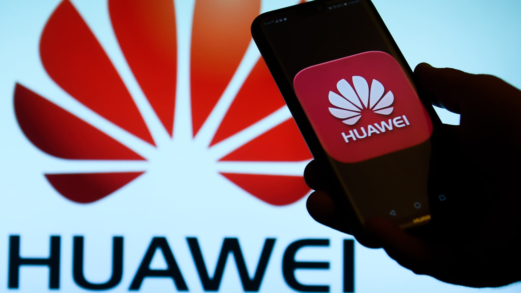 Photographie d'une main tenant un smartphone avec le logo Huawei à l'écran et en fond derrière.