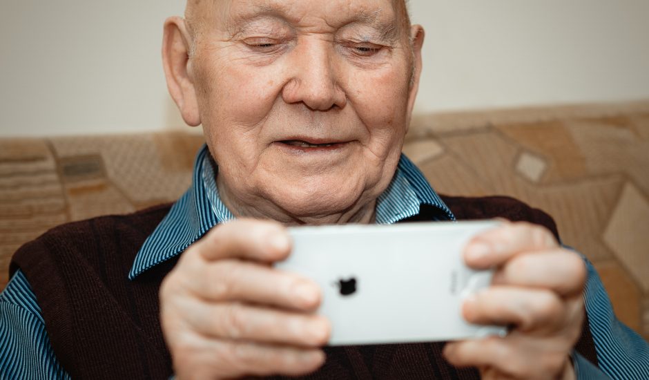 Portrait d'un vieil homme élégant, positif et joyeux, assis sur un divan et tenant dans ses mains un smartphone