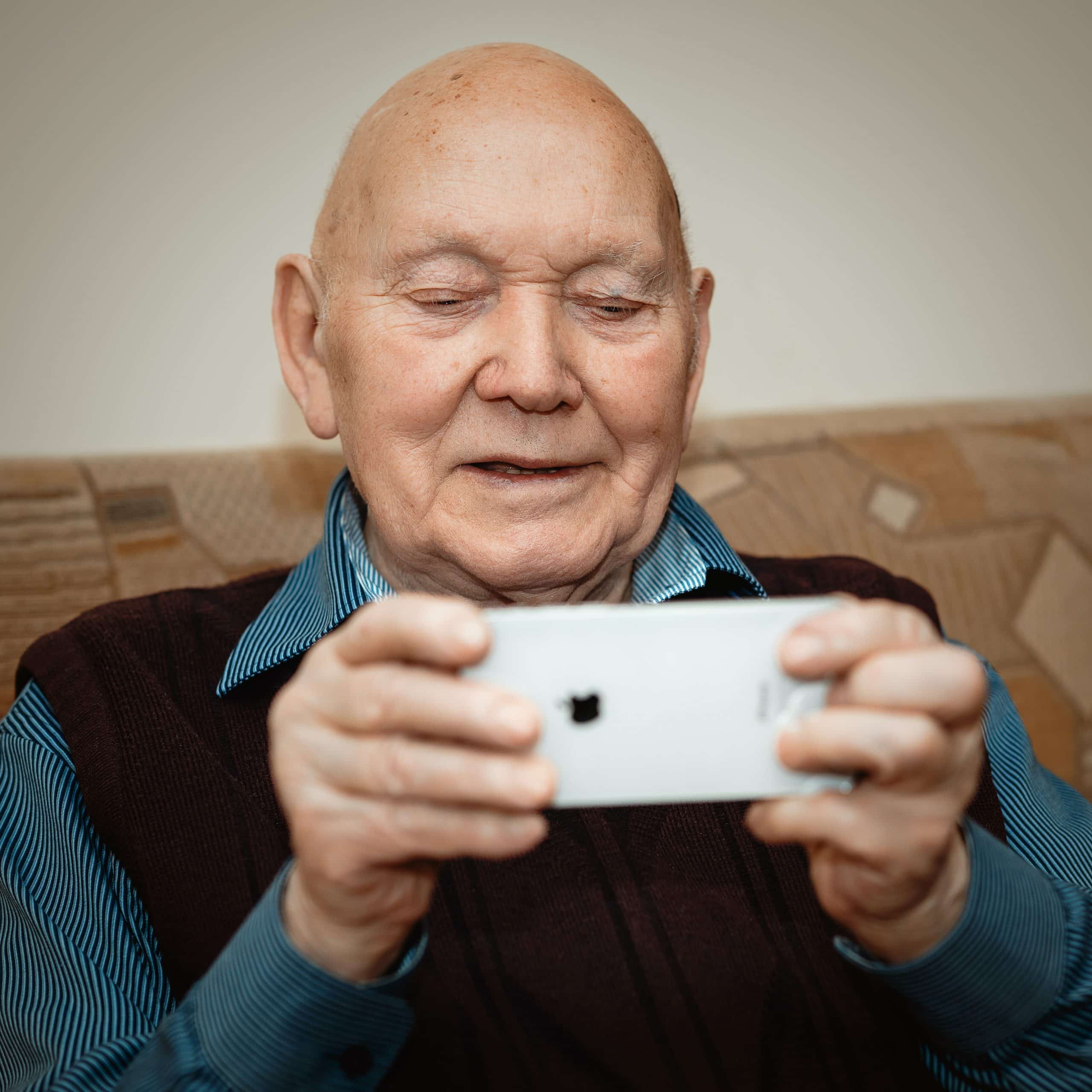 Portrait d'un vieil homme élégant, positif et joyeux, assis sur un divan et tenant dans ses mains un smartphone