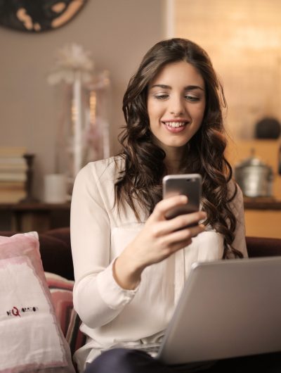 une femme assise sur son canapé souriant devant son téléphone et avec un ordinateur portable sur ses genoux