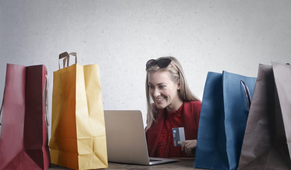Une femme souriant avec sa carte bleue dans les mains devant son ordinateur. Des sacs de couleurs autour
