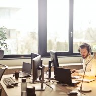 un homme en chemise blanche devant un ordinateur portable et un écran, fenêtre en arrière-plan