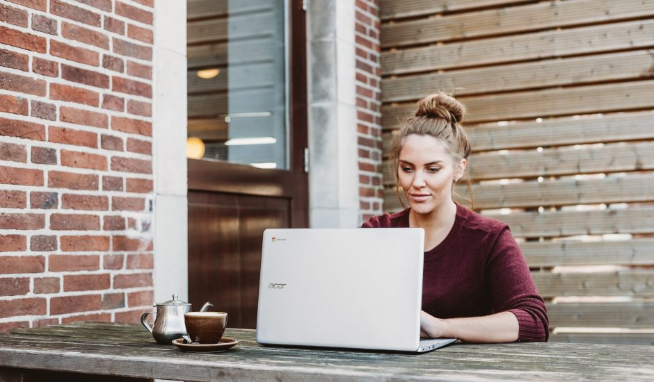 une femme avec un pull bordeaux assise dehors avec un ordinateur portable sur une table en bois
