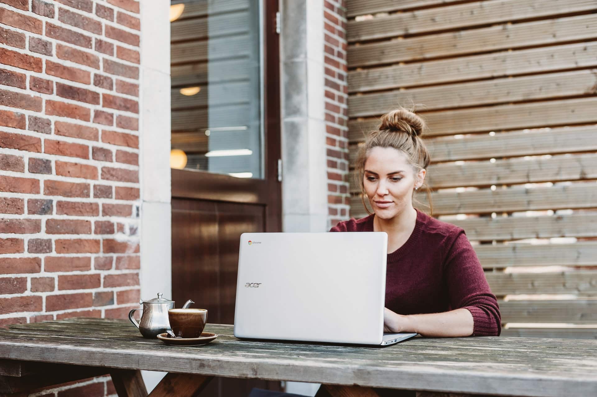 une femme avec un pull bordeaux assise dehors avec un ordinateur portable sur une table en bois