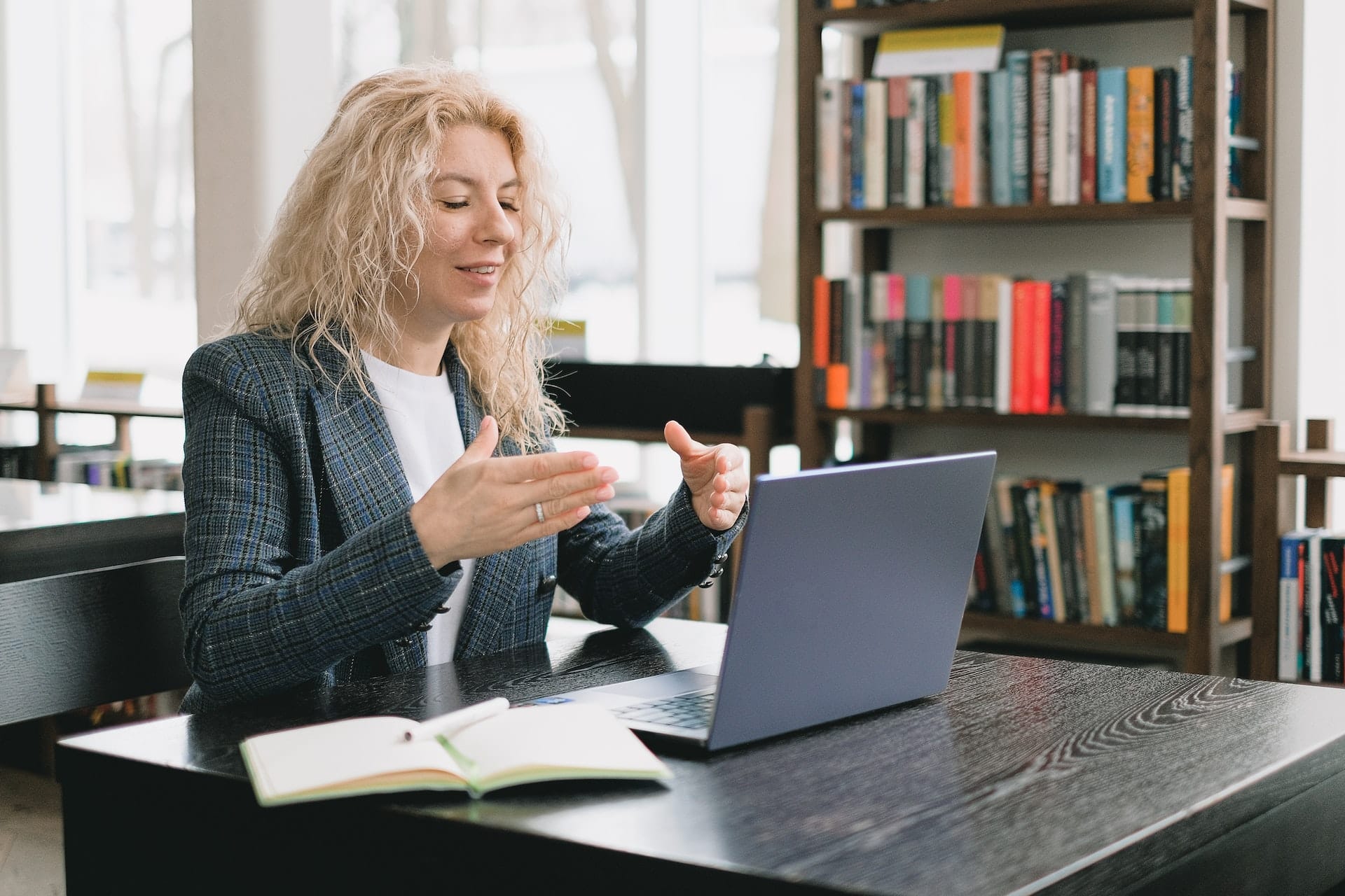 une femme devant un ordinateur portable, carnet sur la table, une bibliothèque en arrière-plan
