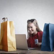 Smiling woman with credit card in hand in front of her computer.  Colorful bags around