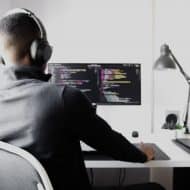un homme de dos, à son bureau, devant un ordinateur en train de coder