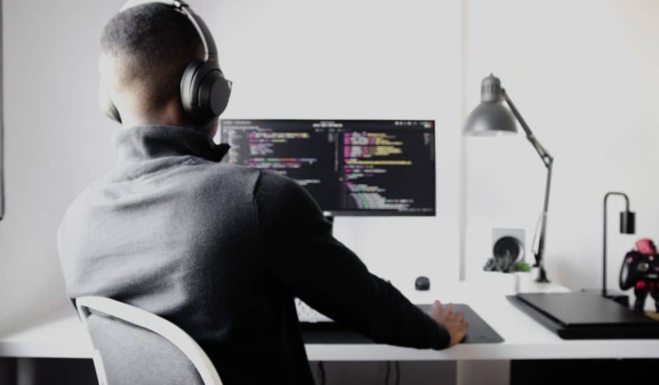 un homme de dos, à son bureau, devant un ordinateur en train de coder