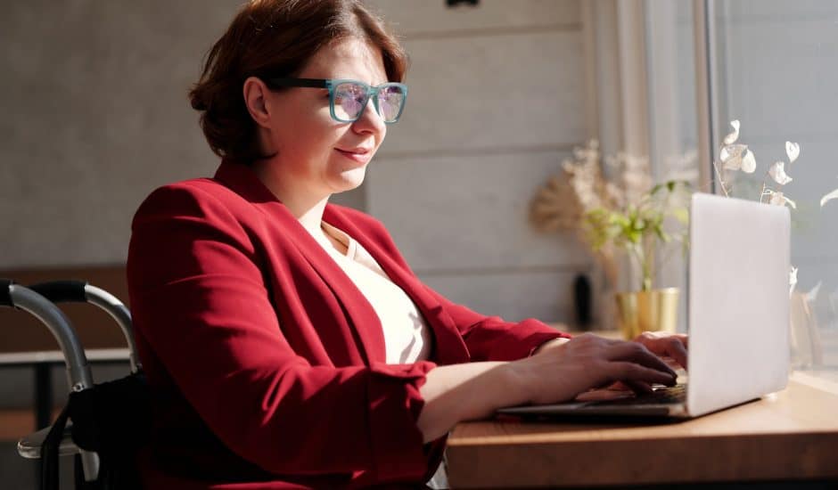 une femme en fauteuil roulant devant son ordinateur portable. Elle a des lunettes et est habillée avec une veste rouge.