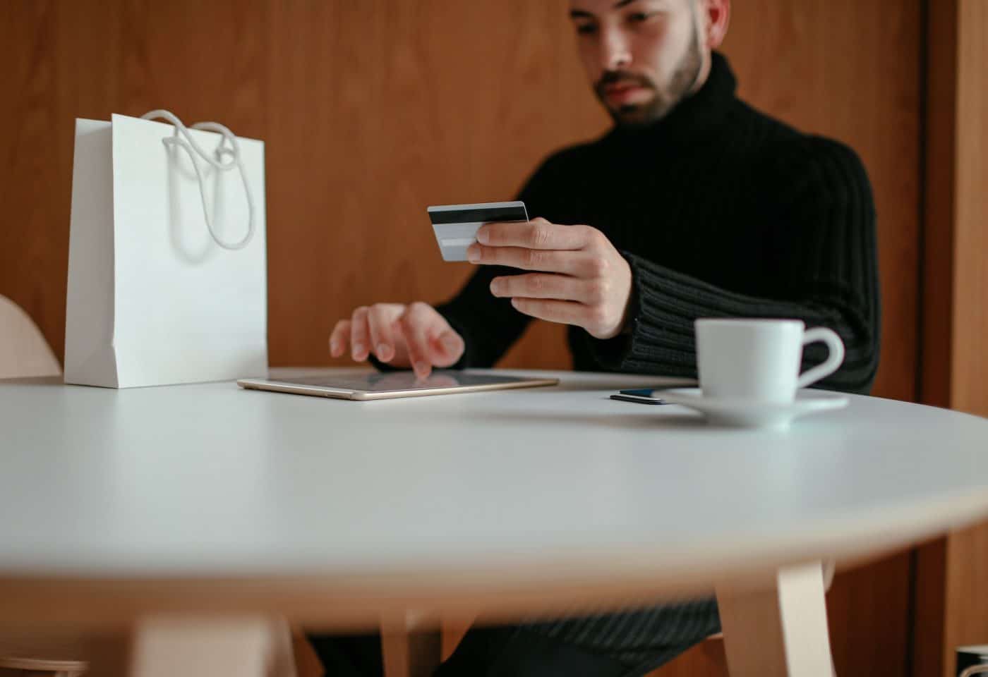 un homme avec une carte bancaire dans la main en train de faire du shopping en ligne. Un sac blanc et une tasse posés sur la table
