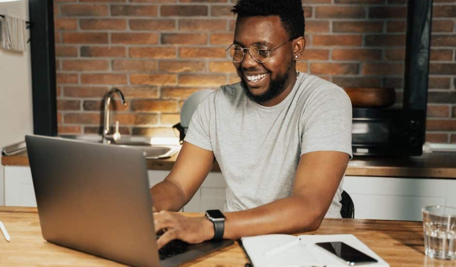 un homme souriant devant un ordinateur, cuisine en arriere-plan