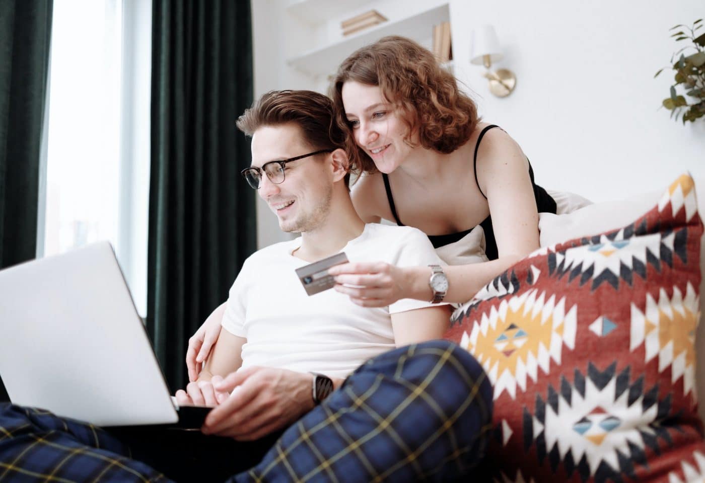 un homme sur le canapé avec l'ordinateur sur les genoux, une femme derrière lui avec une carte bancaire en main