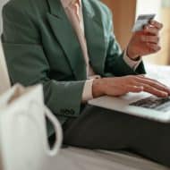 une personne avec une carte bancaire dans la main avec un ordinateur portable gris sur les genoux, sac en papier blanc à sa droite