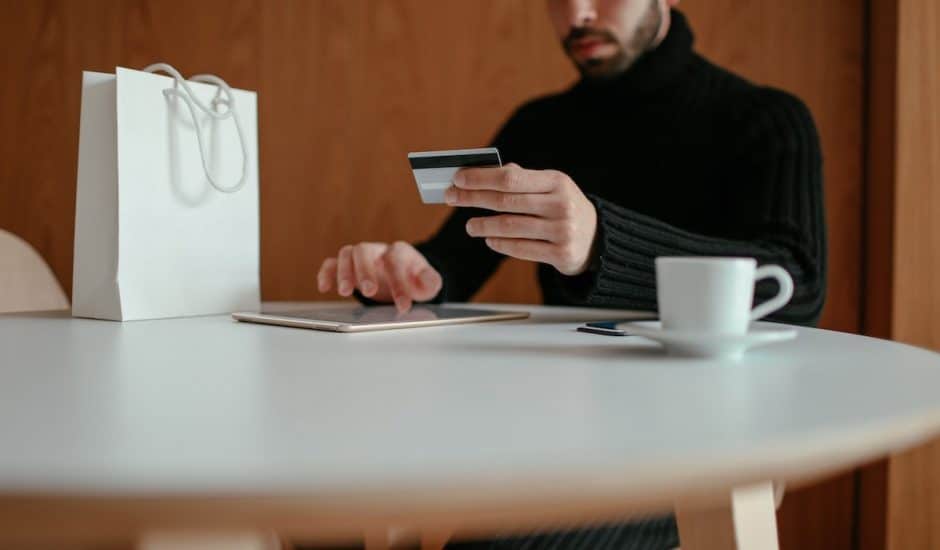 un homme avec une carte bancaire dans la main en train de faire du shopping sur une tablette. Sac blanc et tasse de café sur la table