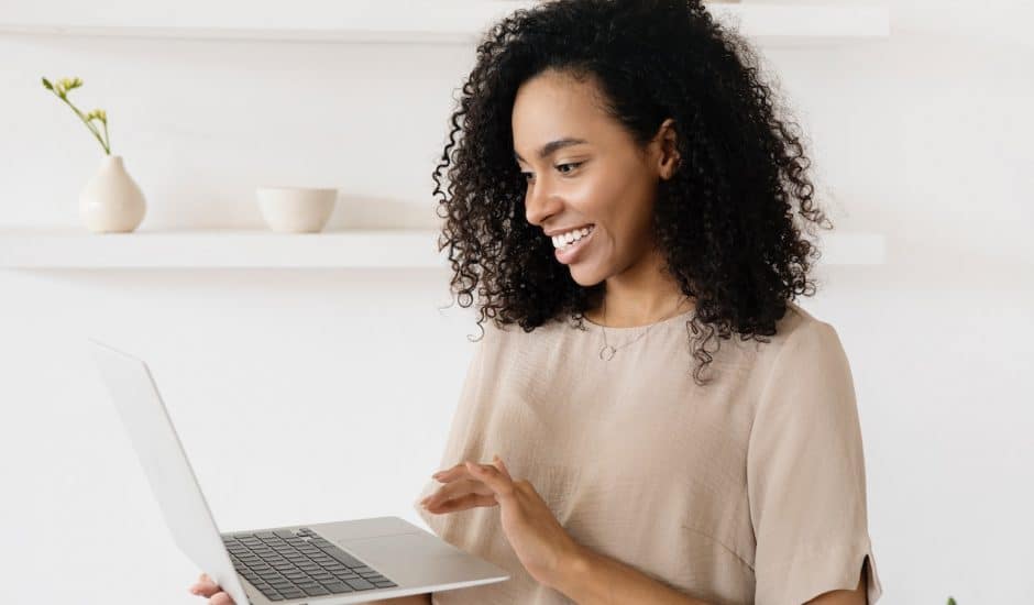 une femme souriante avec un haut marron, elle porte un ordinateur portable dans sa main droite