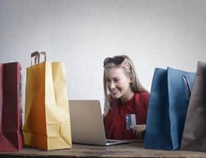 une femme devant avec un ordinateur, carte bancaire en main, des sacs colorés autour d'elle