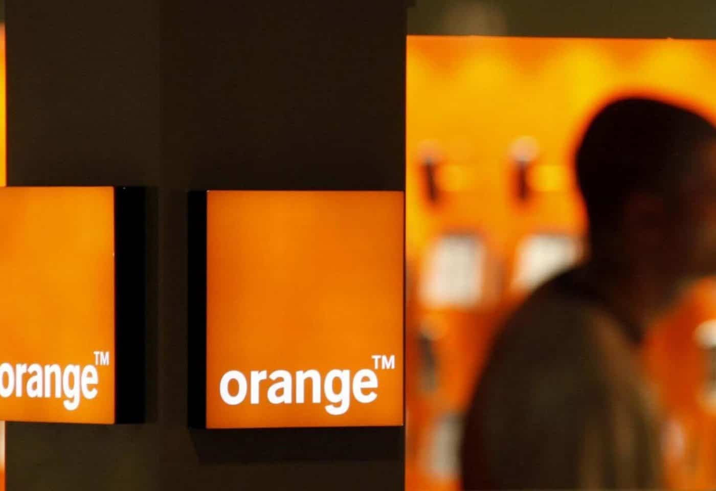 Logo d'Orange sur un poteau, une personne debout en arrière-plan.
