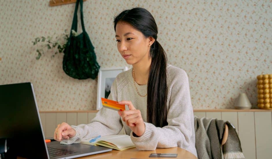 une femme devant son ordinateur avec une carte bancaire orange entre les mains