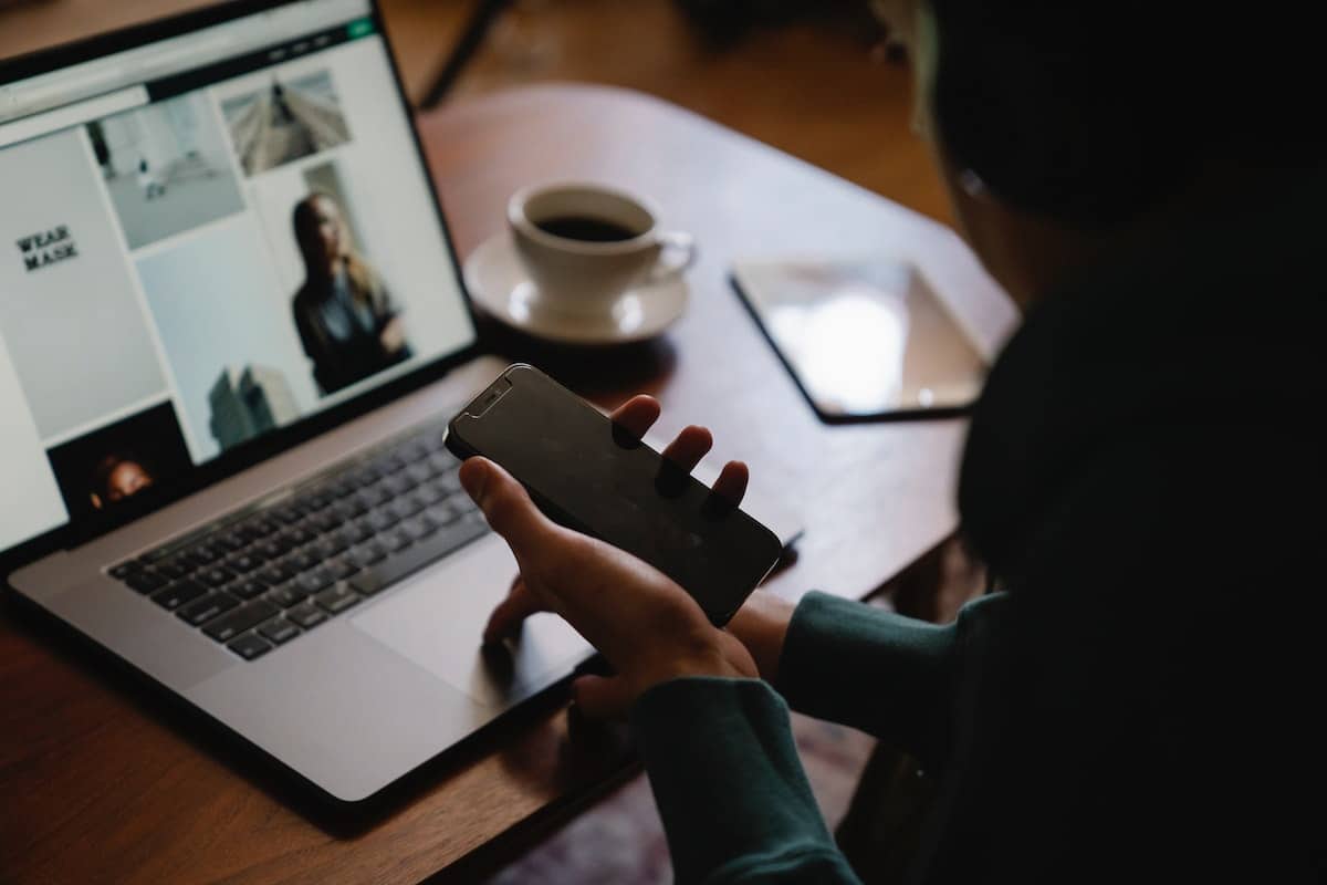 une personne avec un téléphone dans la main, devant un ordinateur avec des visuels dessus. Une tasse de café sur la table en bois