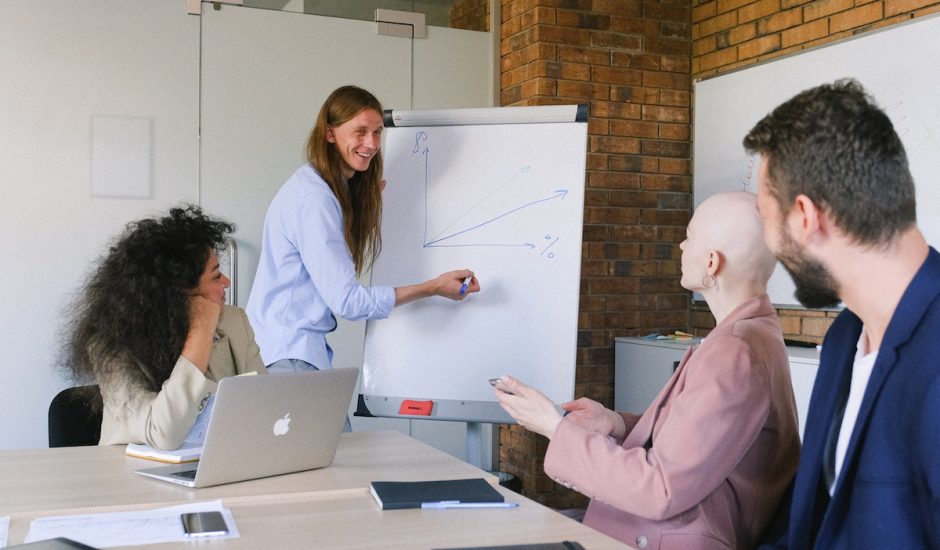 quatre personnes en réunion marketing, une debout devant un tableau avec un graphique et les autres assises le regardant