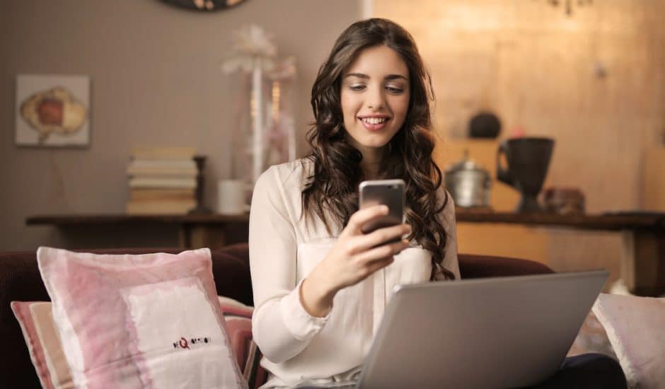 une femme souriante devant son téléphone, ordinateur portable sur les genoux