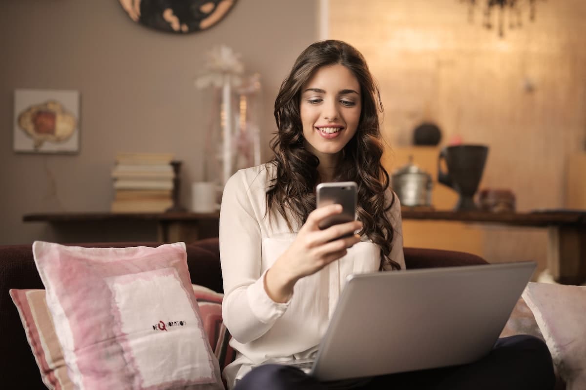 une femme souriante devant son téléphone, ordinateur portable sur les genoux