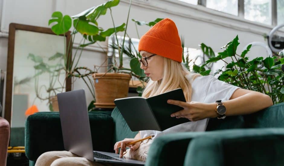 une gemme avec un bonnet orange prenant des notes sur un carnet tout en ayant un ordinateur portable sur les genoux, plantes en arrière-plan