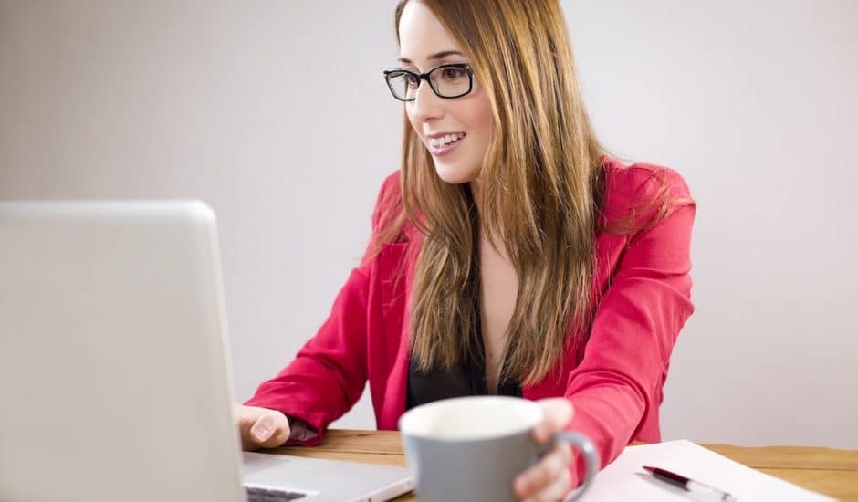 une femme souriante devant son ordinateur, elle tient une tasse de café dans sa main