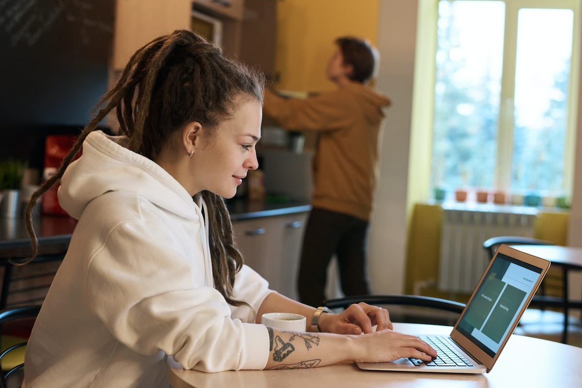 une femme travaillant sur un ordinateur, un homme en fond dans une cuisine