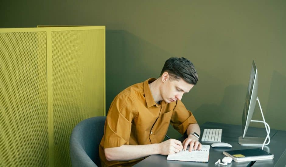 un homme en train d'écrire sur un carnet devant un ordinateur