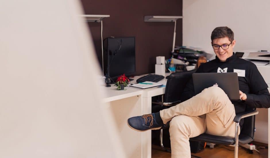 un homme souriant avec son ordinateur portable sur les genoux. Il est dans un bureau