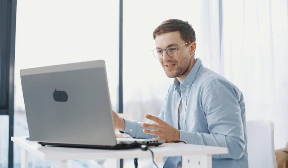 un homme avec des lunettes et une chemise bleue devant un ordinateur portable gris