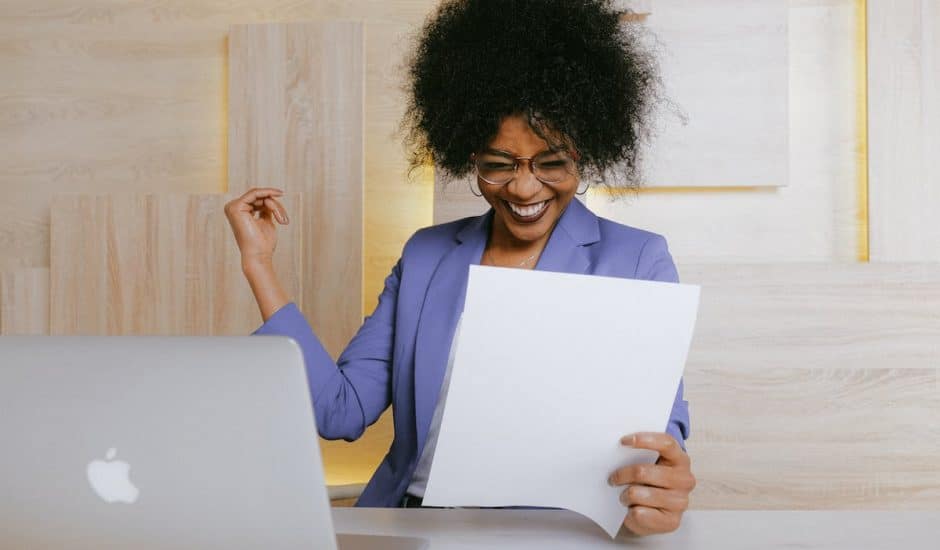 une femme souriante tenant un papier devant son ordinateur