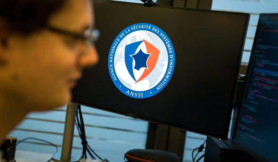 Le logo de l'Anssi sur un écran d'ordinateur au second plan, le visage d'un homme portant des lunettes au premier plan.